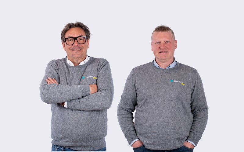 L'image montre un homme souriant, posant dans ce qui semble être un magasin de vélos ou un atelier de réparation. Il porte une chemise polo noire avec le logo "Declerck" brodé sur la poitrine. Il est appuyé contre un comptoir ou une table, avec une roue de vélo visible à côté de lui. Derrière lui, il y a des plantes vertes et un mur bleu, ce qui donne une ambiance accueillante et professionnelle à l'endroit. L'homme semble être un employé ou le propriétaire du magasin.