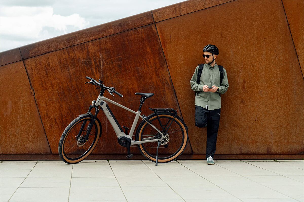 L'image montre un homme debout à côté d'un vélo électrique dans un environnement urbain. L'homme porte un casque de vélo, des lunettes de soleil, une chemise verte et un pantalon noir. Il tient un téléphone portable dans sa main droite, comme s'il était en train de vérifier quelque chose ou d'attendre un appel. Le vélo est un vélo électrique, identifiable par la batterie intégrée dans le cadre. Le vélo est stationné sur un sol pavé devant un mur métallique rouillé, ce qui donne un contraste intéressant entre le vélo moderne et le mur industriel. Le ciel est partiellement nuageux, ce qui suggère une journée avec un temps variable.