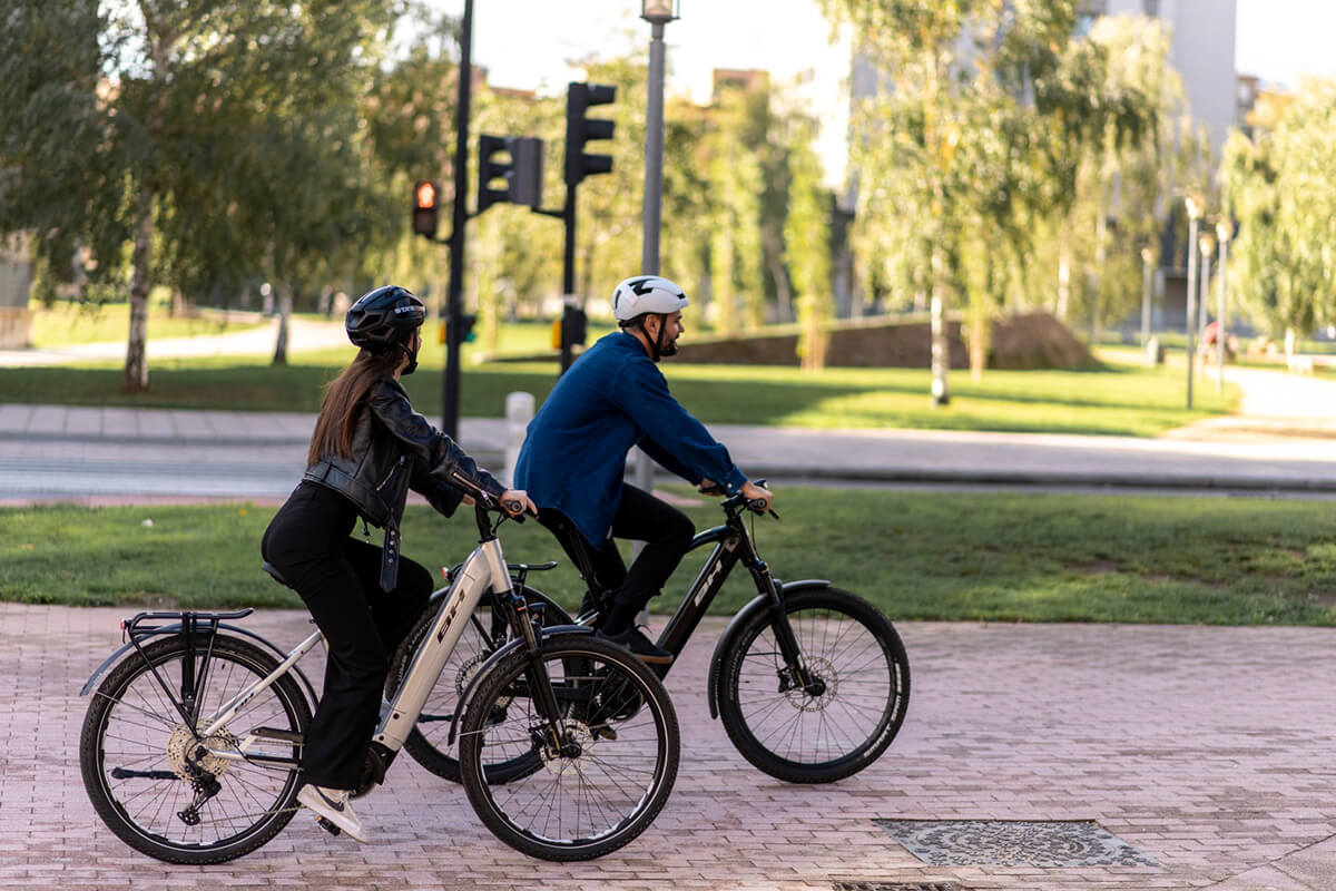 L'image montre deux personnes à vélo dans un environnement urbain. Les deux cyclistes portent des casques de sécurité. La personne à gauche porte une veste noire et un pantalon noir, tandis que la personne à droite porte un sweat-shirt bleu et un pantalon noir. Ils sont tous les deux sur des vélos électriques, identifiables par la batterie visible sur le cadre des vélos. Ils semblent attendre à un feu de circulation, qui est visible en rouge à l'arrière-plan. Le cadre est un espace vert avec des arbres et des pelouses, et il y a un trottoir pavé sur lequel ils se trouvent. Le ciel est clair, suggérant une journée ensoleillée.
