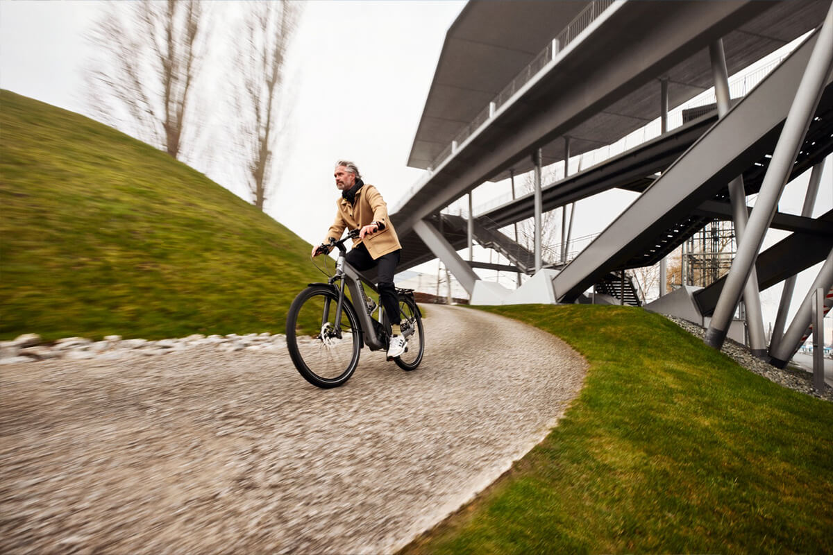 L'image montre une personne à vélo sur un chemin en gravier sous un pont moderne. La personne porte une veste beige et un pantalon noir, et elle semble se déplacer à une vitesse modérée, ce qui est suggéré par le flou de mouvement autour d'elle et du vélo. Le vélo est un vélo électrique, identifiable par la batterie visible sur le cadre. Le chemin est bordé de gazon bien entretenu et mène sous une structure de pont métallique complexe, ce qui donne une impression de modernité et d'infrastructure urbaine. En arrière-plan, on peut voir des arbres sans feuilles, indiquant que la saison pourrait être l'automne ou l'hiver. Le ciel est couvert, ce qui ajoute une ambiance fraîche à la scène.
