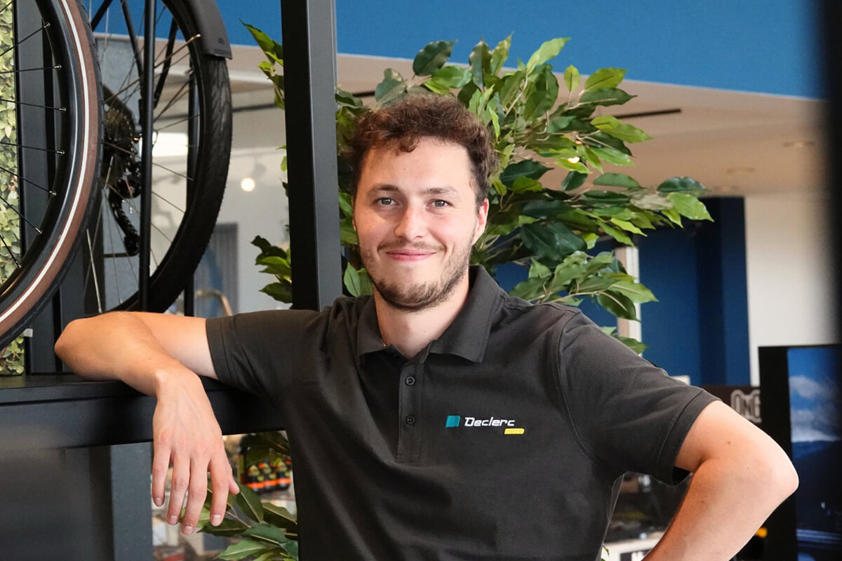 L'image montre un homme souriant, posant dans ce qui semble être un magasin de vélos ou un atelier de réparation. Il porte une chemise polo noire avec le logo "Declerck" brodé sur la poitrine. Il est appuyé contre un comptoir ou une table, avec une roue de vélo visible à côté de lui. Derrière lui, il y a des plantes vertes et un mur bleu, ce qui donne une ambiance accueillante et professionnelle à l'endroit. L'homme semble être un employé ou le propriétaire du magasin.