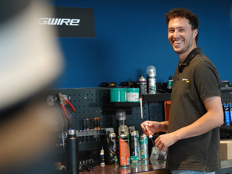 L'image montre un homme souriant dans ce qui semble être un atelier ou un magasin de réparation. Il porte une chemise polo avec un logo et se tient devant un mur bleu où le mot "GWIRE" est affiché. Sur le comptoir devant lui, il y a divers outils et produits, notamment des bombes aérosols, des pinces et d'autres équipements de réparation. L'homme semble être en train de travailler ou de préparer quelque chose, et l'environnement suggère qu'il pourrait s'agir d'un atelier de réparation de vélos ou d'un magasin spécialisé dans les équipements de sport ou de plein air.